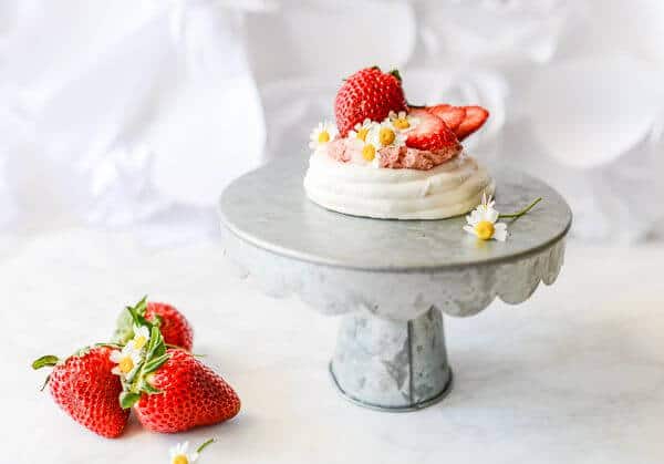 A small strawberry Pavlova is placed on a cake plate. Three strawberries are in the foreground.