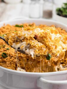 A close up of a spoonful of warm cheesy chicken hash brown casserole being lifted from a white casserole dish.