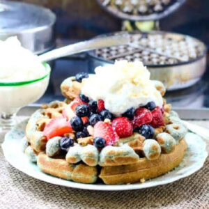 Blue cornmeal waffles with strawberries and blueberries on top on a plate.