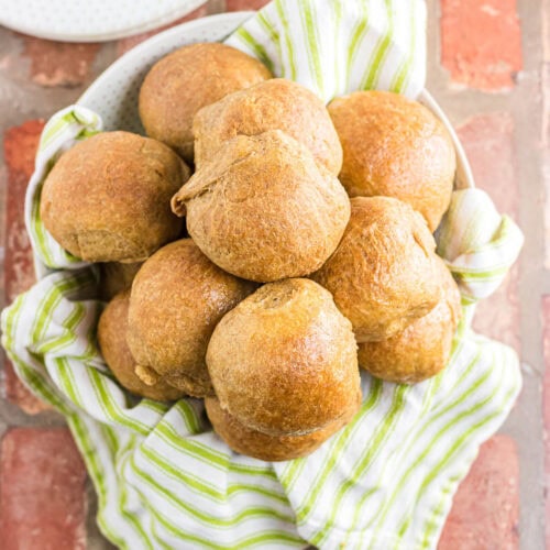 A bowl of whole wheat dinner rolls with a green and white striped towel.