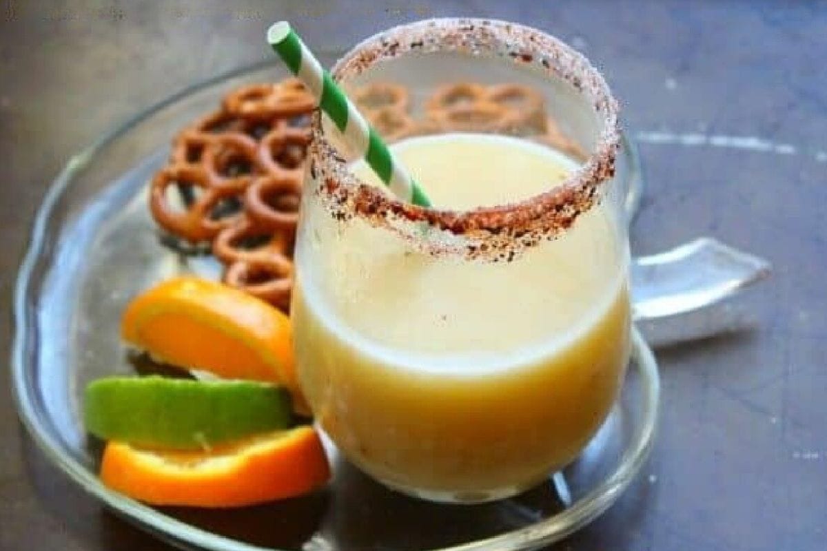 A frozen banana margarita on a table with a plate of pretzels and fruit.