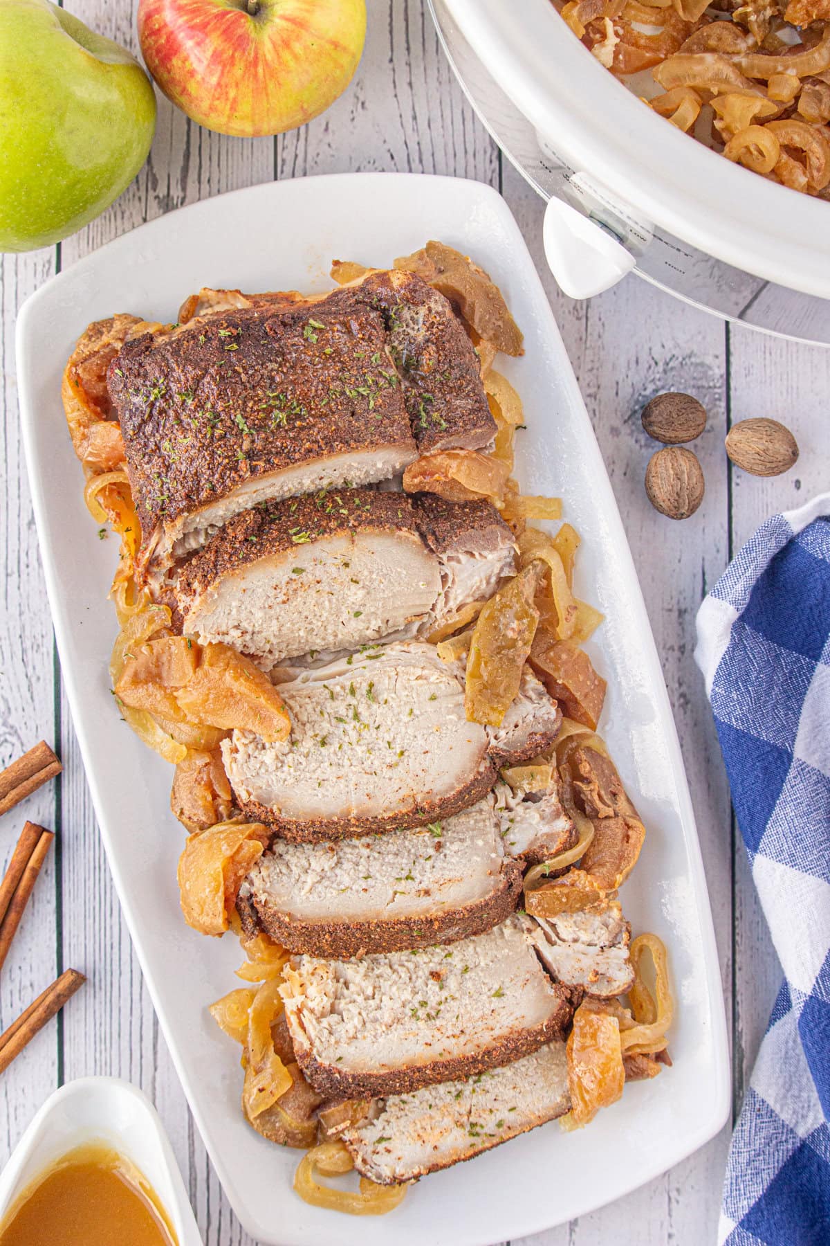 Sliced pork loin on a platter with the apple and onion slices, next to the slow cooker off to the side.