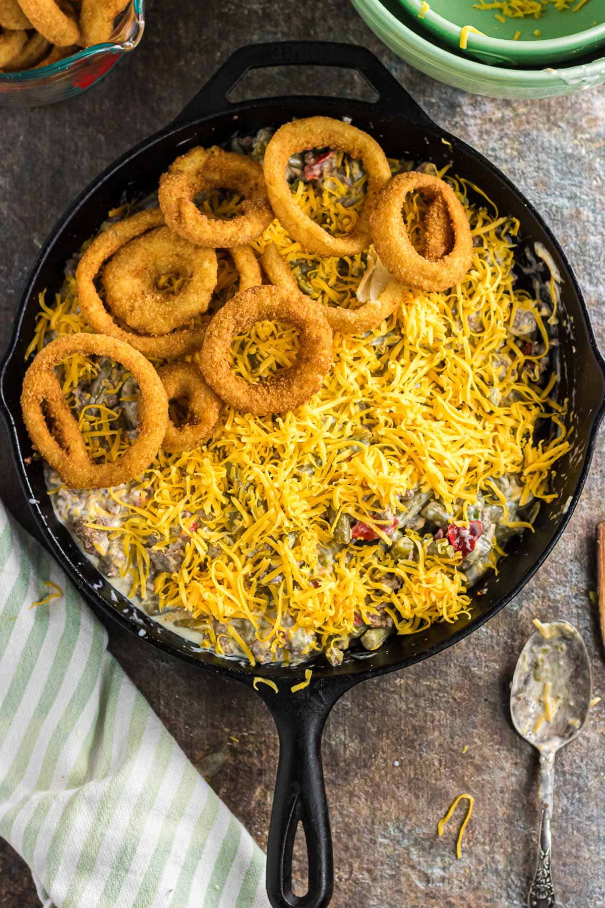 Adding cheese and onion rings on top of the green beans and ground beef in the skillet.