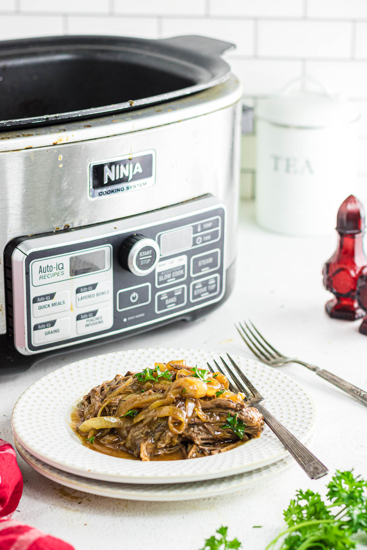 A serving plate of French onion pot roast next to a fork and slow cooker.