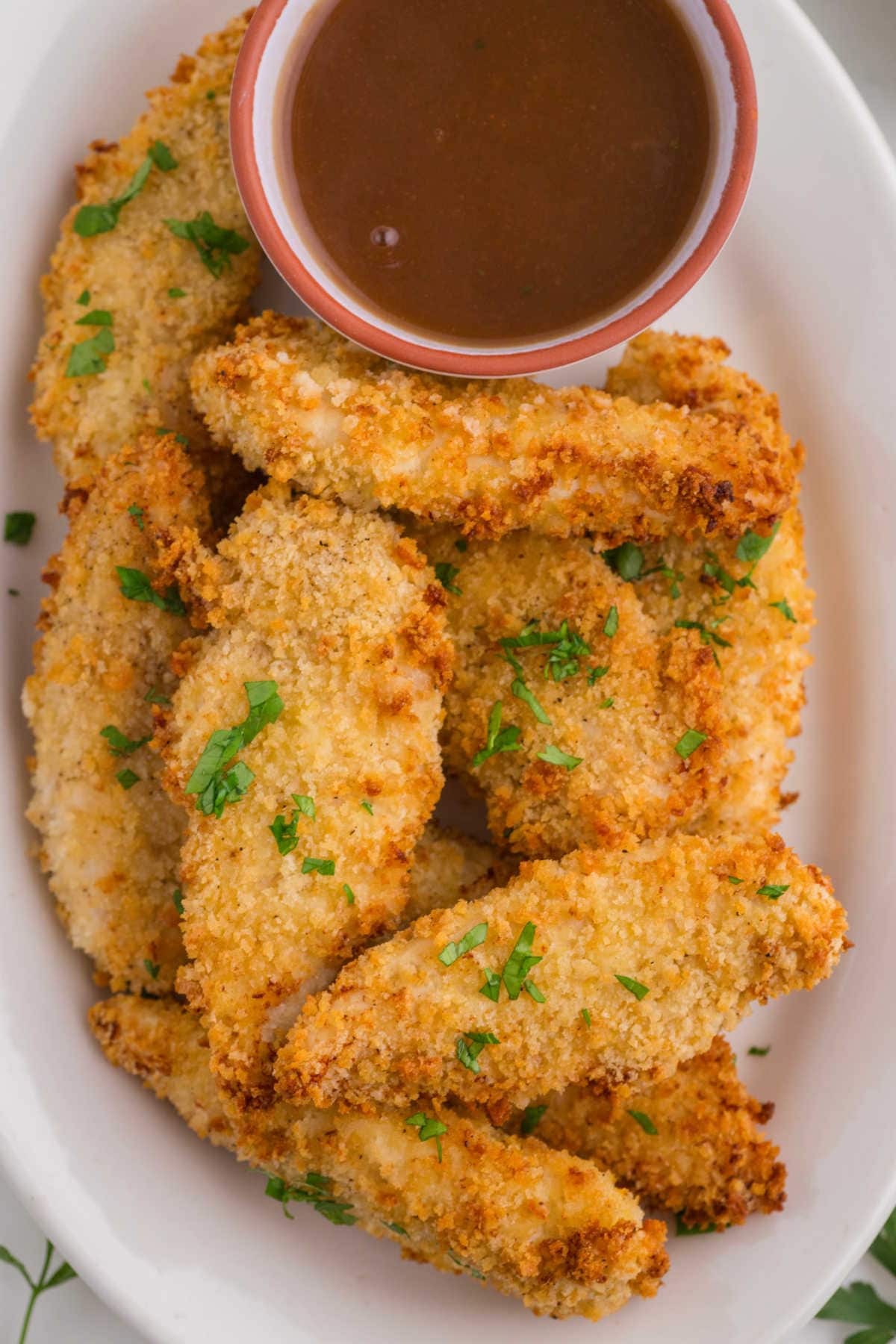 Overhead view of crispy chicken tenders on a white plate with a small bowl of dipping sauce.