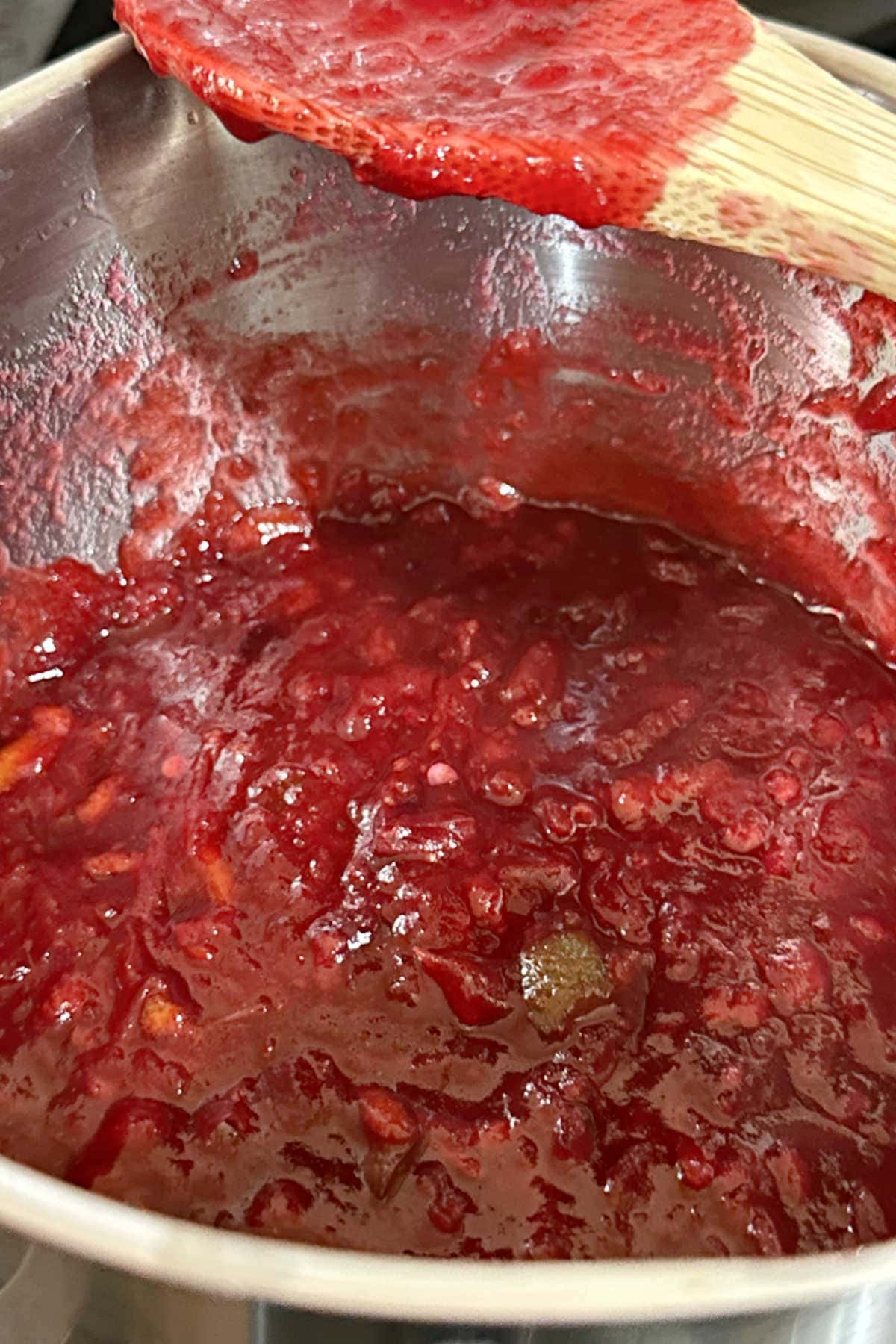 The cranberries have popped open and the sauce is shown thickened on a wooden spoon.