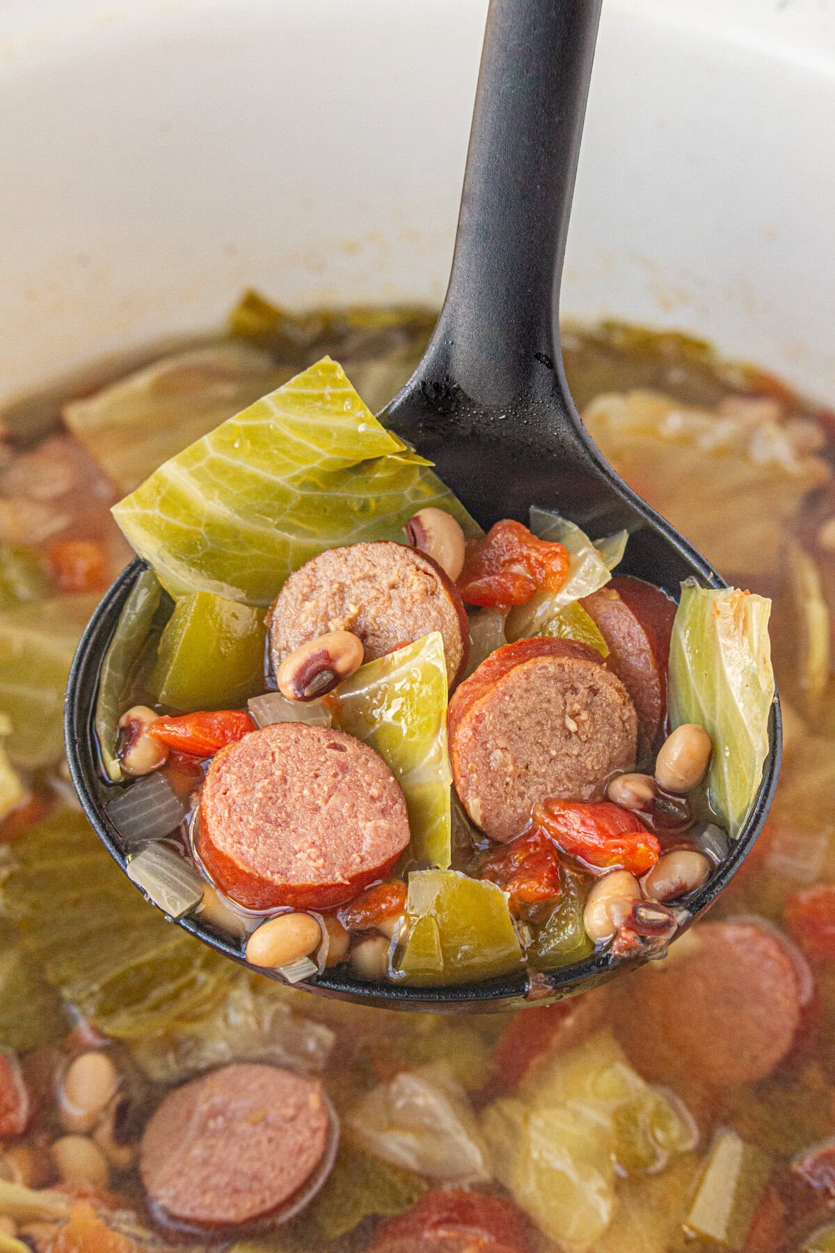 An up-close view of sausage, black eyed pea, and cabbage soup in a ladle.