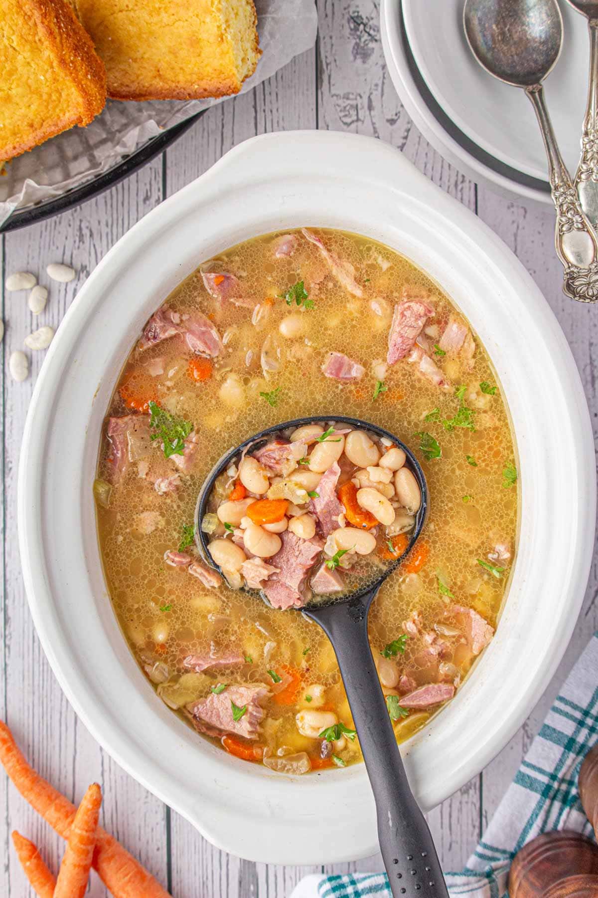 An overhead view of a ladle scooping slow cooker ham and bean soup from the crockpot.