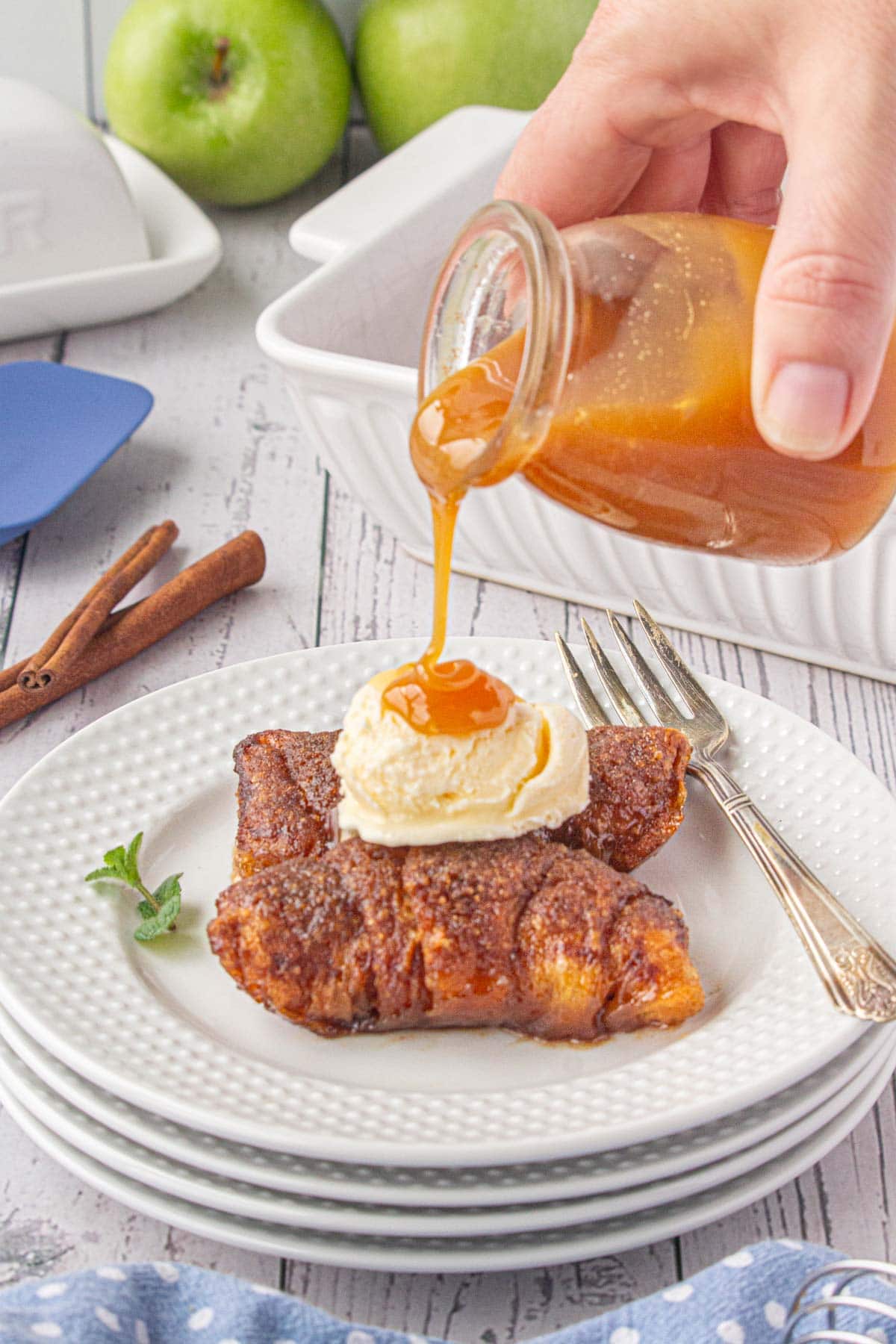 Pouring caramel sauce over crescent roll apple dumplings topped with a scoop of ice cream.